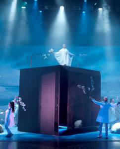 Stora scenen i blått ljus, skådespelare utspridda dansande. Strålkastare fårn taket, en kvinna i vitt i mitten på en stor svart kub