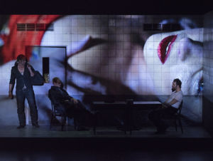 Föreställningbild från Carmen, två män sitter vid ett bord, en tredje man står bredvid och håller i en väska.