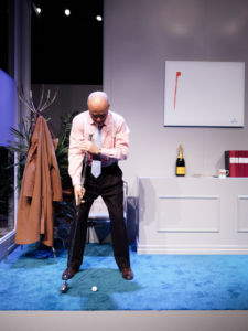 Skådespelaren Gustav Levin spelar golf i tjusiga kläder (repetition på lilla scenen). Blå matta, kontorsmiljö.