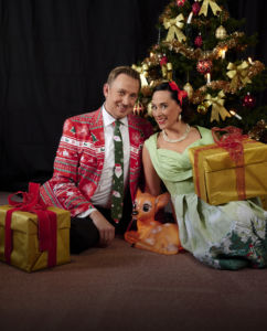 Skådespelarna Daniel Engman och Åsa Forsblad Morisse platstigt leende mot kameran framför en julgran i julkläder