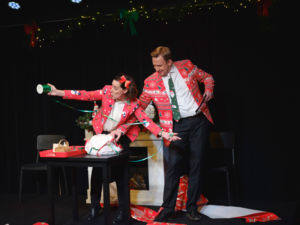 Skådespelarna Åsa Forsblad Morisse och Daniel Engman slår in paket på scenen i röda julkavajer