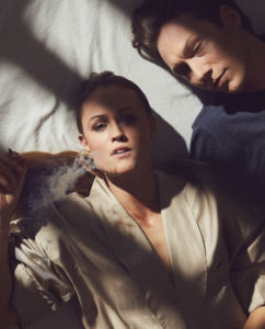 Kvinna och man i morgonljus i en säng taget ovanifrån. Kvinnan röker och tittar in i kameran. Mannen tittar på henne.