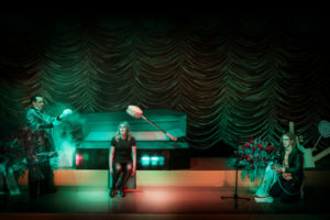 En skådespelare framför en öppen kista, med en undersökningslampa ovanför sitt huvud. Grönt ljus på scenen
