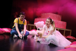 två stycken ungdomar leker med barbiedockor på golvet, rosa nallebjörn sitter med
