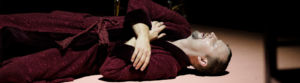 Skådespelaren Aksel Morisse som Doktor Glas, ligger på golvet i röd morgonrock och tar sig för bröstet
