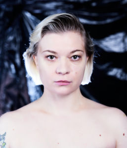 Porträttfoto Rita Lemivaara mot en bakgrund av svart sopsäck, nakna axlar