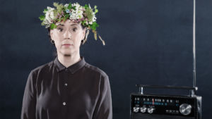 SKådespelaren Åsa Forsblad Morisse med en blomsterkrans på huvudet sittanades vid ett bord, en gammaldags radio bredvid henne på bordet