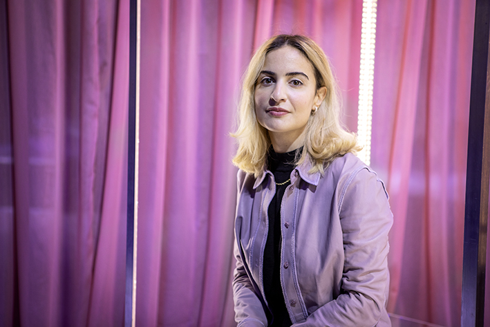 En kvinna i lila skjorta sitter framför ett rosa tyg