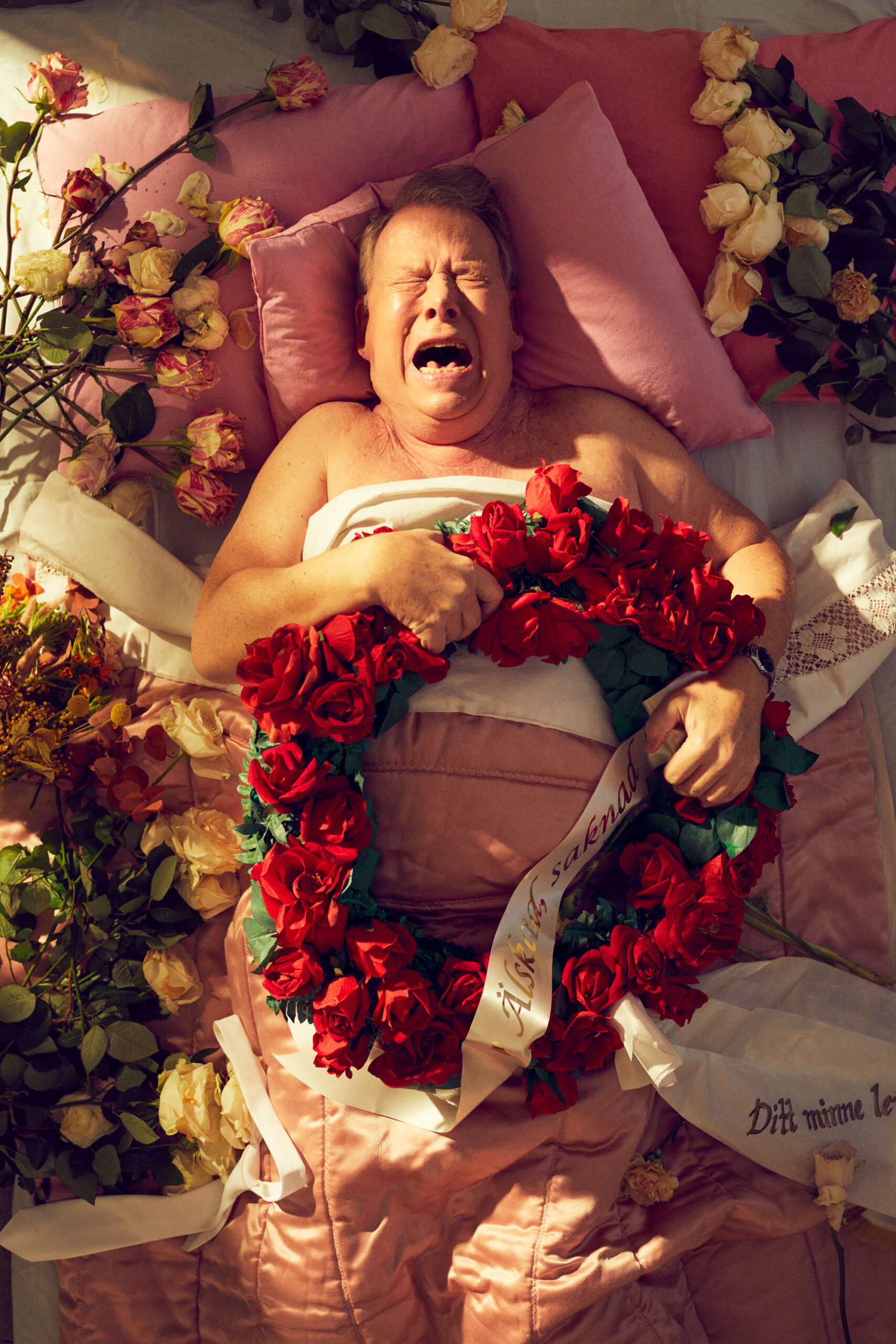 Man som ligger i en säng och vrålar av sorg, blommor över sig