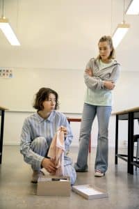 två unga kvinnor titta på varandra i ett klassrum, en sitter ner och öppnar en låda, den andre står upp