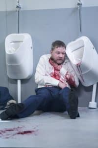 En man sitter på golvet vid en pissoar. Har blod på kläderna och händerna