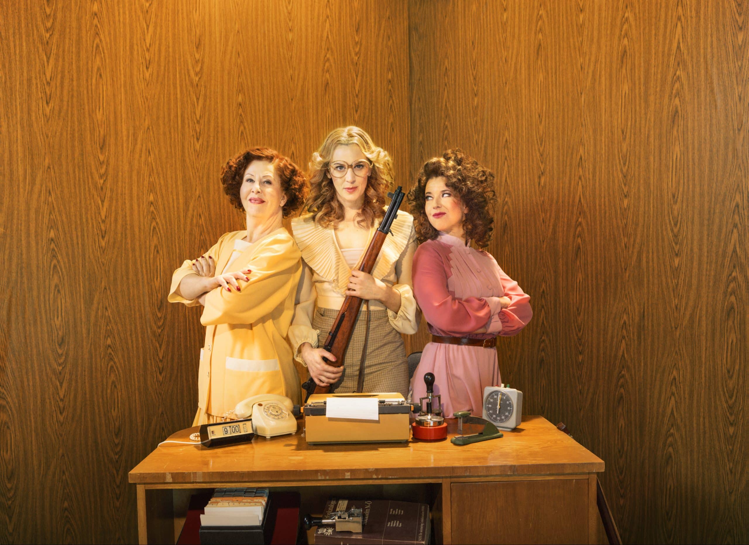 Tre kvinnor iklädda kläder från 70-talet i kontorsmiljö. Kvinnan i mitten håller ett gevär