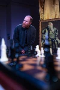 En man står hukad bredvid ett porträtt av Gustav Vasa, i förgrunden syns ett schackbord