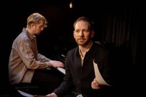 två män på en scen, en vid piano och en med papper i handen
