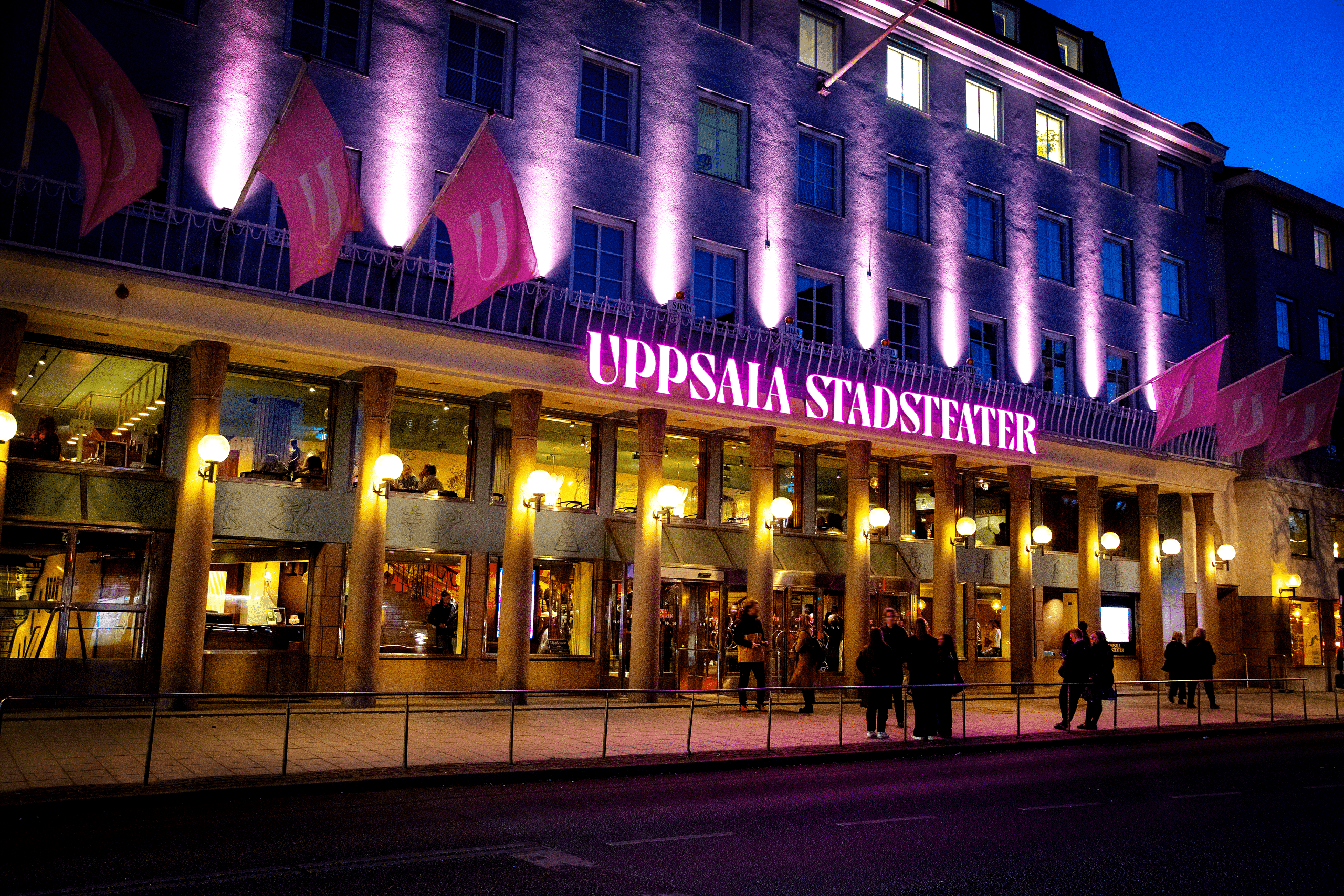 en fasad i mörker med rosa lampor riktade mot taket och en rosa neonskylt där det står Uppsala stadsteater